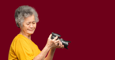 Happy senior woman looking at her photos in a digital camera while standing on a red background. Aged people photographer and relaxation concept. Space for textの写真素材