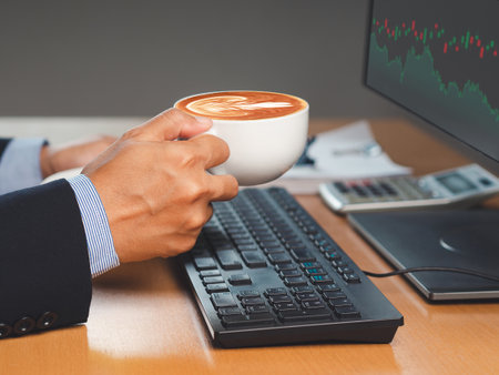 Hand of a businessman holding a coffee cup and looking at a graph stock on a screen monitor while sitting in the office. Close-up photo. Business, finance, and investment conceptの写真素材