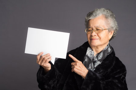 Speech bubble concept. An elderly Asian woman in a sweater with short gray hair holds and points to a blank speech bubble with a smile while standing on a gray backgroundの写真素材