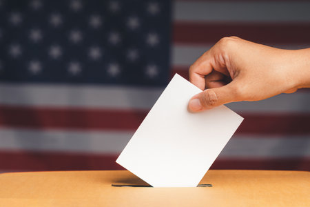 Hand voter holding ballot paper putting into the voting box at place election with an American flag...