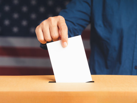 Hand voter holding ballot paper putting into the voting box at place election with an American flag...