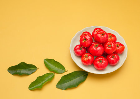 Top view of ripe red acerola cherries on a white dish and green leaves over a yellow background....