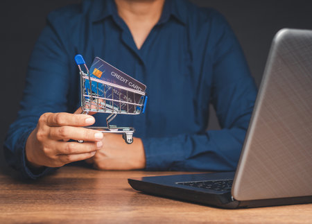 Man in a blue shirt holding a mini shopping trolley with a card mockup while sitting at the table in the office. Concept of online shopping. Close-up photoの写真素材