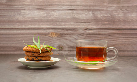 Cannabis chocolate chip cookies on a plate with a marijuana leaf and a glass of cannabis tea with wooden wall background. Concept of cooking with cannabis herbの写真素材
