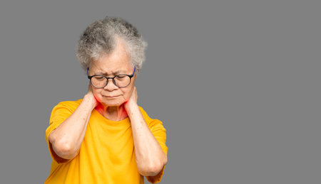 Portrait of a senior woman suffering neck pain while standing on a gray background. Aged people and...