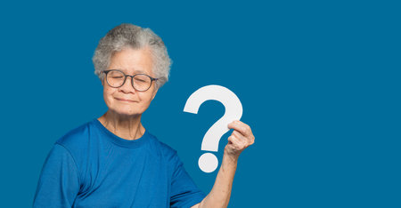 Question mark concept. An elderly Asian woman with short white hair holding a white question mark paper with a smile while standing on a blue background. Space for textの写真素材