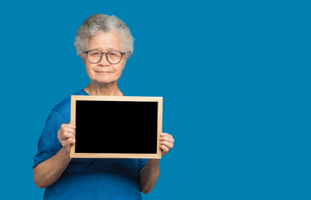 Portrait of an elderly Asian woman with short gray hair holding a mini blackboard and looking at the camera with a smile while standing on a blue background. Aged people and relaxation conceptの写真素材