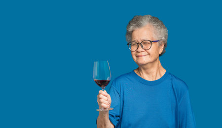Portrait of a senior woman with short white hair holding a red wine glass and looking at the camera with a smile while standing on a blue background. Aged people, Beverage and relaxation conceptの写真素材