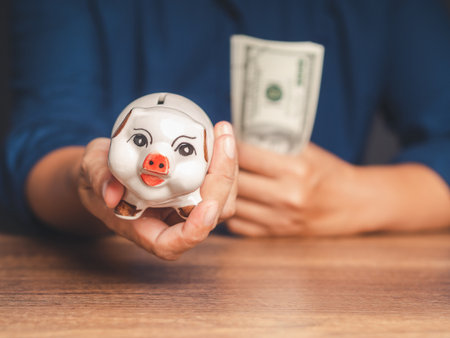 Close-up of hand holding of a piggy bank and US dollar bills while sitting at the table. Money and savings concept. Selective focusの写真素材
