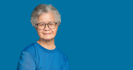 Portrait of an elderly Asian woman with short gray hair looking at the camera with a smile while standing on a blue background in the studio. Space for text. Aged people and relaxation conceptの写真素材
