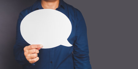 Speech bubble concept. Close-up of a hand businessman in a blue shirt holding a blank white speech bubble while standing on a gray background. Space for textの写真素材