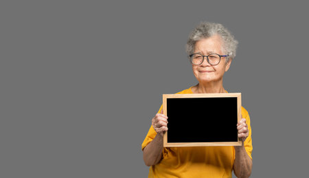 Portrait of an elderly Asian woman with short gray hair holding a mini blackboard and looking at the camera with a smile while standing on a gray background. Aged people and relaxation conceptの写真素材