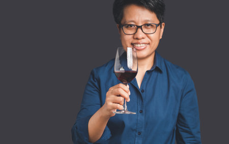 Portrait of a woman with short black hair in a blue shirt holding a red wine glass while standing on a gray background. Space for text. Beverage and relaxation conceptの写真素材
