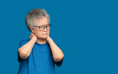 Portrait of a senior woman suffering neck pain while standing on a blue background. Aged people and...