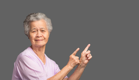 Portrait of an elderly Asian woman with short gray hair pointing with two hands and fingers to the...