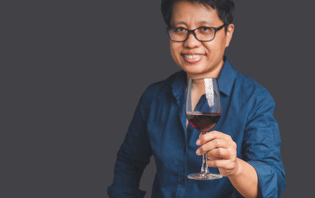 Portrait of a woman with short black hair in a blue shirt holding a red wine glass while standing on a gray background. Space for text. Beverage and relaxation conceptの写真素材
