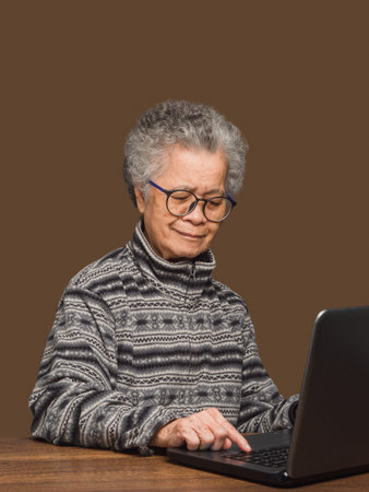 An elderly Asian woman with short grey hair uses a laptop to search for data from the internet while...
