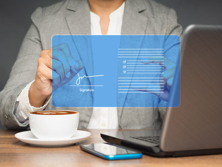 A businesswoman in a suit uses a pen to sign electronic documents on a virtual screen while sitting at the table in the office. Technology, document management, and paperless office conceptの写真素材