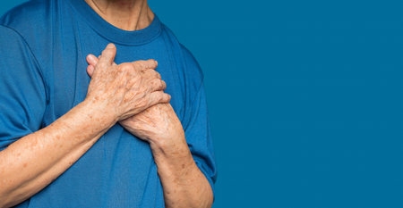 A senior woman in blue casual has sore left chest pain while standing on a blue background. Close-up photo. Space for text. Aged people and the healthcare conceptの写真素材