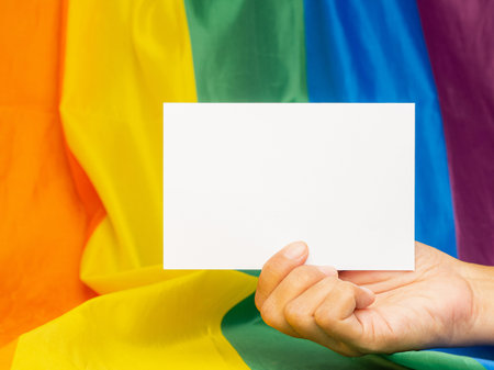 Hand holding a blank white paper against the rainbow flag or LGBT flag background. Space for text. Gender diversity concept. Close-up photoの写真素材
