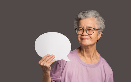 Speech bubble concept. An elderly Asian woman with short white hair holding a blank speech bubble and looks at the camera with a smile while standing on a gray backgroundの写真素材