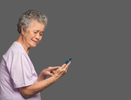 An elderly Asian woman is using a smartphone with a smile while standing on a gray background. Aged people and communication conceptの写真素材