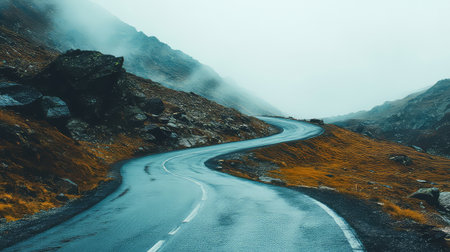 A winding road snakes through a foggy mountain terrain, showcasing a tranquil journey amidst breathtaking scenery. The wet asphalt contrasts with dry, rugged slopes, making the landscape a captivating sight for nature lovers and travelers.の素材