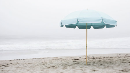 A tranquil beach setting featuring a light blue umbrella perched on soft sand, with gentle ocean waves lapping in the background under a cloudy sky.の素材