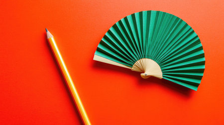 A vibrant flat lay image featuring a green paper fan and a yellow pencil on a bright orange background, perfect for artistic and creative projects.の素材
