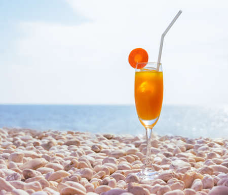 Orange juice with carrot slice in cocktail glass on white pebbly beach with seascape background, summer tropical holiday concept. Vintage filtered image.の写真素材