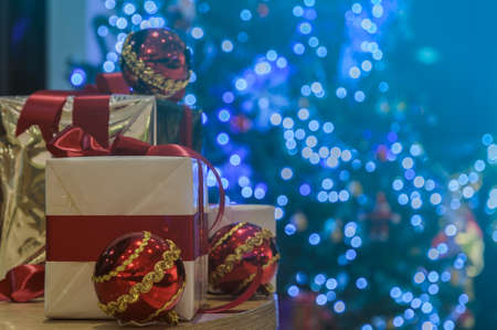 Gift boxes with red bow and red balls on table with illuminated Christmas tree