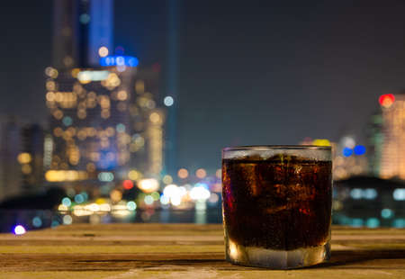 Close up of a glass of  carbonated soft drink or cola with ice and night view of bokeh urban city lightsの写真素材