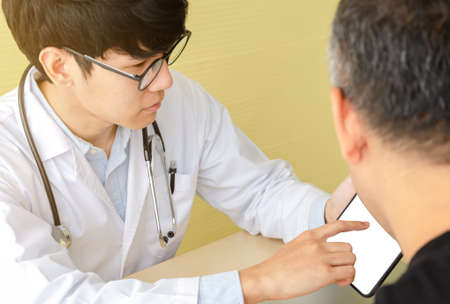 Young asian doctor with glasses talking with male patient by using of blank white screen digital tablet for medical history or laboratory test result. Healthcare and medical with technology conceptの写真素材