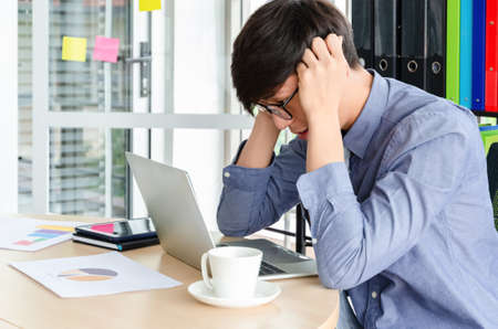 Stressed young Asian businessman sitting at desk looking at labtop and touching his temples with...