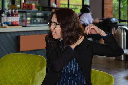 Painful Asian woman wearing glasses on couch working in cafe has shoulder pain or stiffness after...