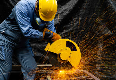 Worker man in blue uniform with yellow satety helmet and leather gloves cutting metal pipe by electric circular abrasive saw with sparks. Industrial working with personnel protective equipment concept.の写真素材