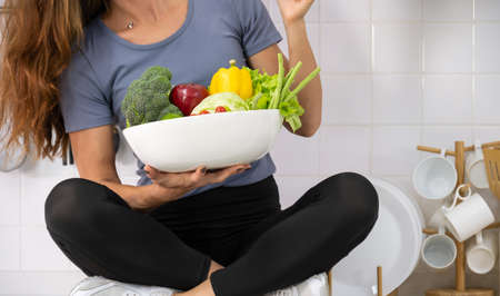 Young woman sitting on kitchen cabinet counter with fresh vegetable. Woman in sportswear with white bowl of  fresh mixed vegetables in her hand. Healthy food and dieting concept.の写真素材