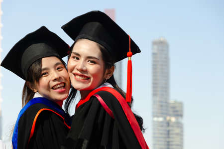 Two happy female in graduation gowns hugging and smiling while  standing outdoors with city buildings backgroundの写真素材