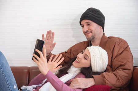Happy couple greeting in a phone video call while relaxing on sofa in living room at homeの写真素材