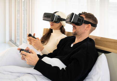 Smiling couple with virtual reality goggles and playing video games with controller in bed. Gaming and technology conceptの写真素材