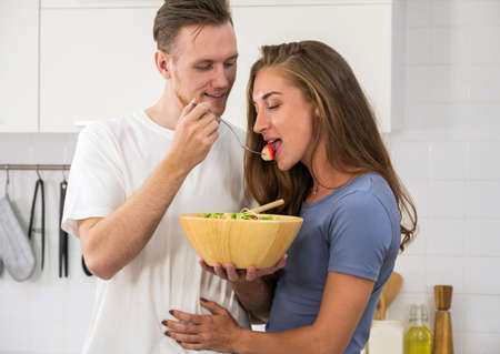 Happy young couple enjoying and preparing a healthy meal in home kitchen. Man holding salad bowl and offering a strawberry to womanの写真素材