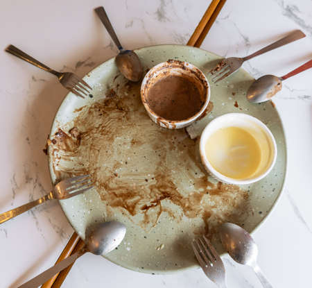 Empty plate with food leftovers from dessert meal, spoons and forks on marble table. Dirty plate with chocolate sauce, chocolate and vanilla ice cream leftovers in ramekin bowl.の写真素材