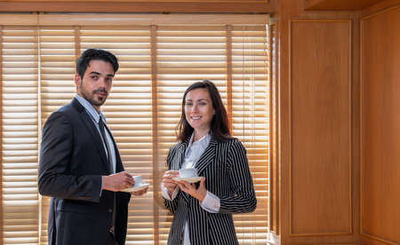 Young businesswoman and  businessman wearing suits and having cups of coffee while standing next to the blind window in office. Business meetings conceptの写真素材