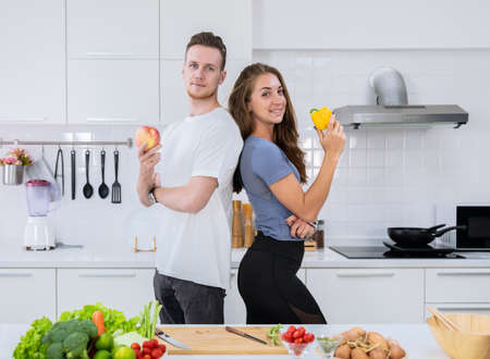 Smiling couple standing in kitchen while preparing healthy food.  Man holding red apple and woman holding yellow bell pepper.の写真素材