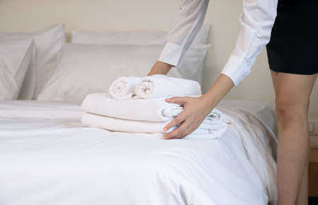 Hotel room service making room. Woman chambermaid keeping stack of fresh white bath towels on bed in guest roomの写真素材