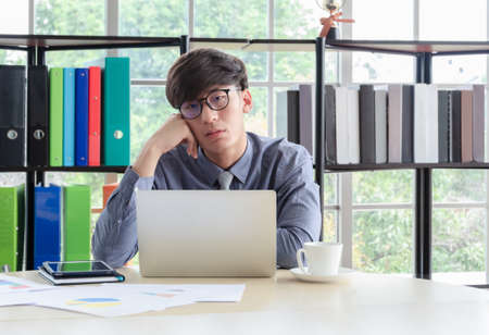 Young sleepy businessman wearing glasses sitting and holding head on hand in front of laptop computer at office desk with boried and tired face expressionの写真素材