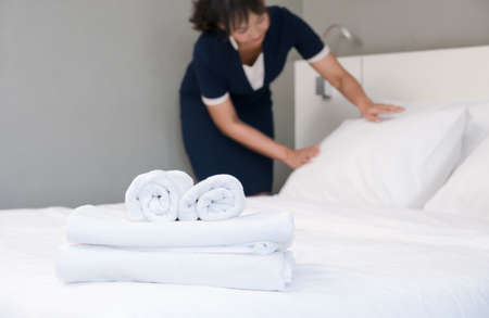 Clean white towels on bed with young hotel maid making bed on backgroundの写真素材