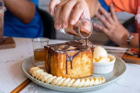 Honey toast served with sliced banana, vanilla and chocolate ice cream on white marble table. Woman hand pouring chocolate sauce on Honey Toastの写真素材