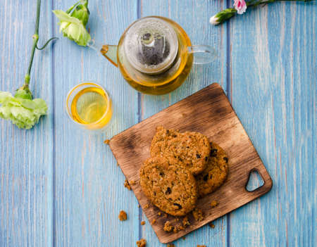 Homemade wholegrain cookies with raisin on wooden cutting board, a cup of tea, teapot and fresh flowers on blue wooden table, top view.の写真素材