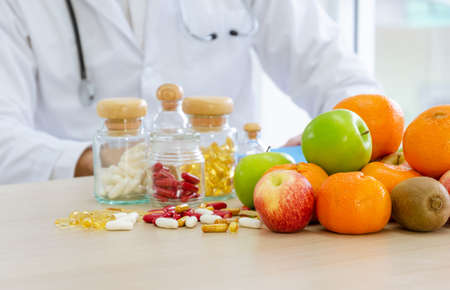 Fresh fruits, capsules and vitamins on desk.  Nutritional expert with stethoscope around neck developing dietary plan to improve eating habits of patientの写真素材